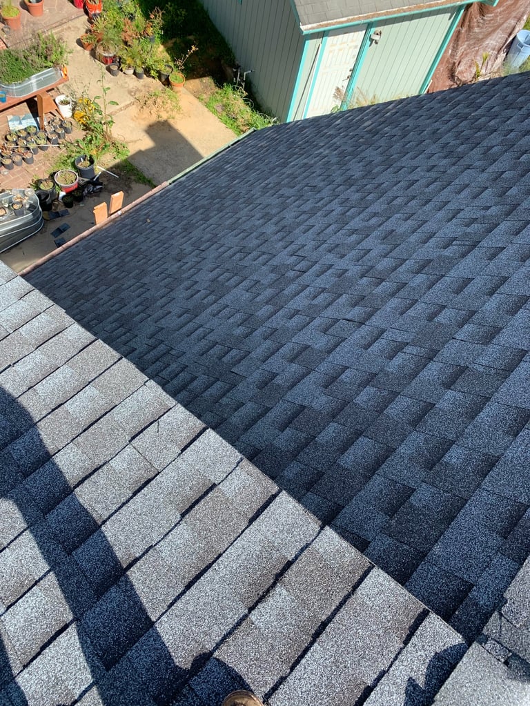 Aerial view of a roof with newly installed asphalt shingles in gray and dark blue tones, adjacent to a metal warehouse building and construction site below