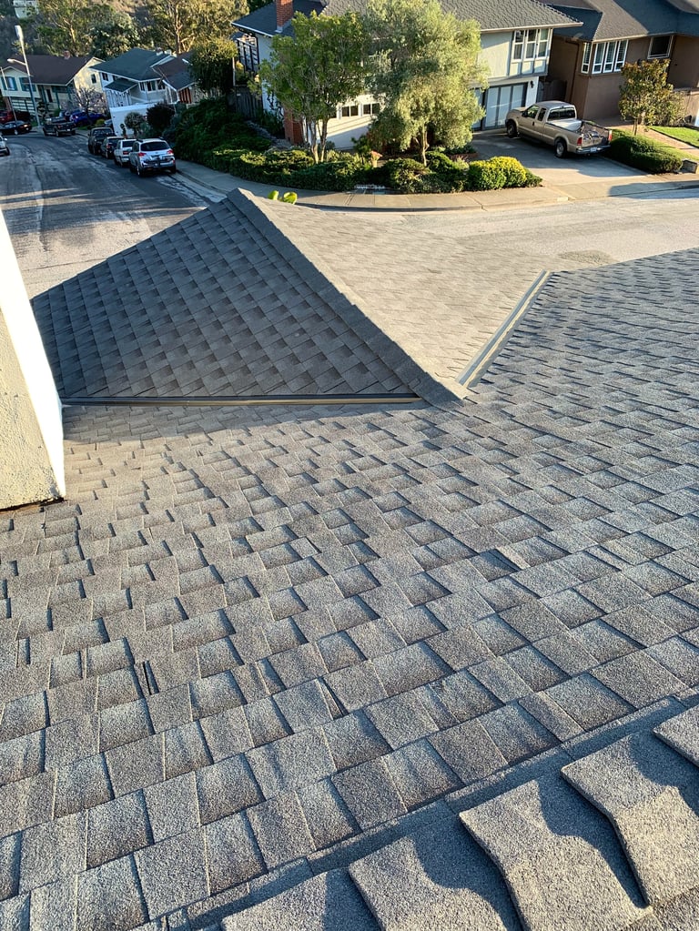 Aerial view of residential roofing with dark and light gray shingles, showing driveway, green landscaping, and neighboring houses