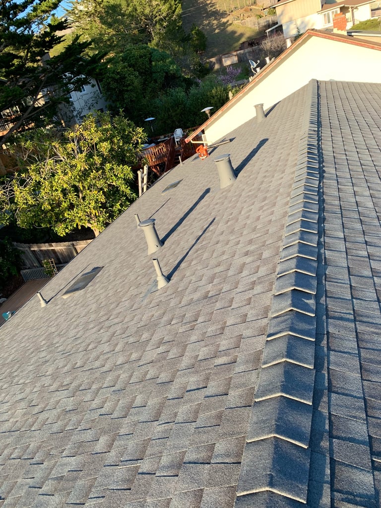 Angled rooftop view showing gray asphalt shingles, metal flashing, and shadows cast by dormers on a residential roof installation