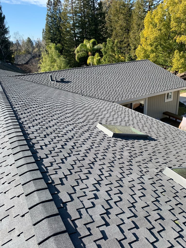 Newly installed gray asphalt shingle roof on a house with forested background and tall trees