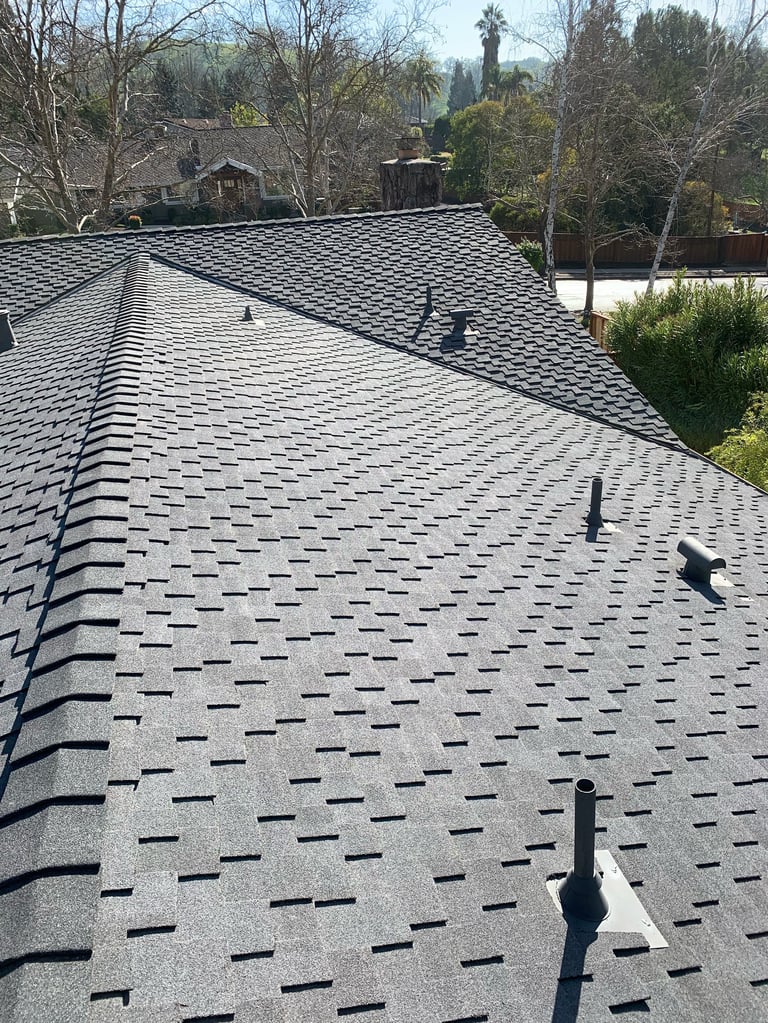Newly installed dark asphalt shingle roof with ventilation pipes on residential home surrounded by trees