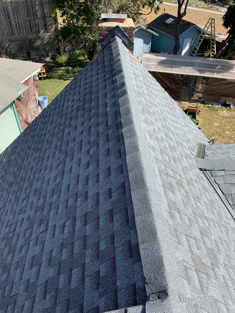 Aerial view of a dark gray asphalt shingle roof with ridge line, overlooking residential yards and structures